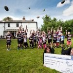 Photograph by Enigma Communications of children throwing rugby balls in the air and man receiving giant cheque in foreground.