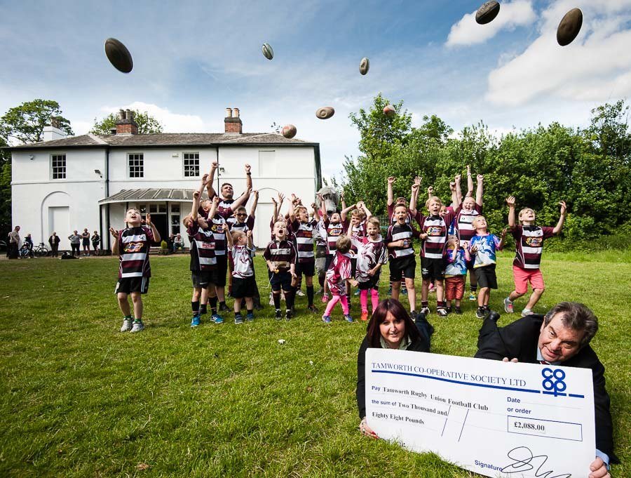 Photograph by Enigma Communications of children throwing rugby balls in the air and man receiving giant cheque in foreground.